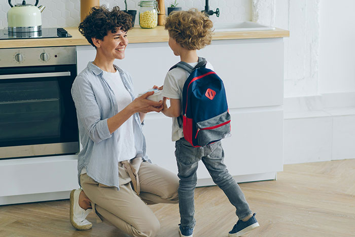 Woman smiling at young boy with backpack in kitchen, illustrating reasons men got upset at women.