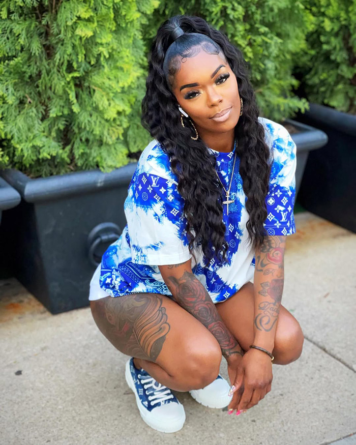 Woman with long curly hair and tattoos wearing a blue and white shirt crouching outside near greenery and black planters.