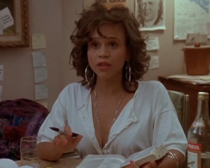 Rosie Perez sitting at a table with a book, discussing Woody Harrelson's comment about her body during filming.