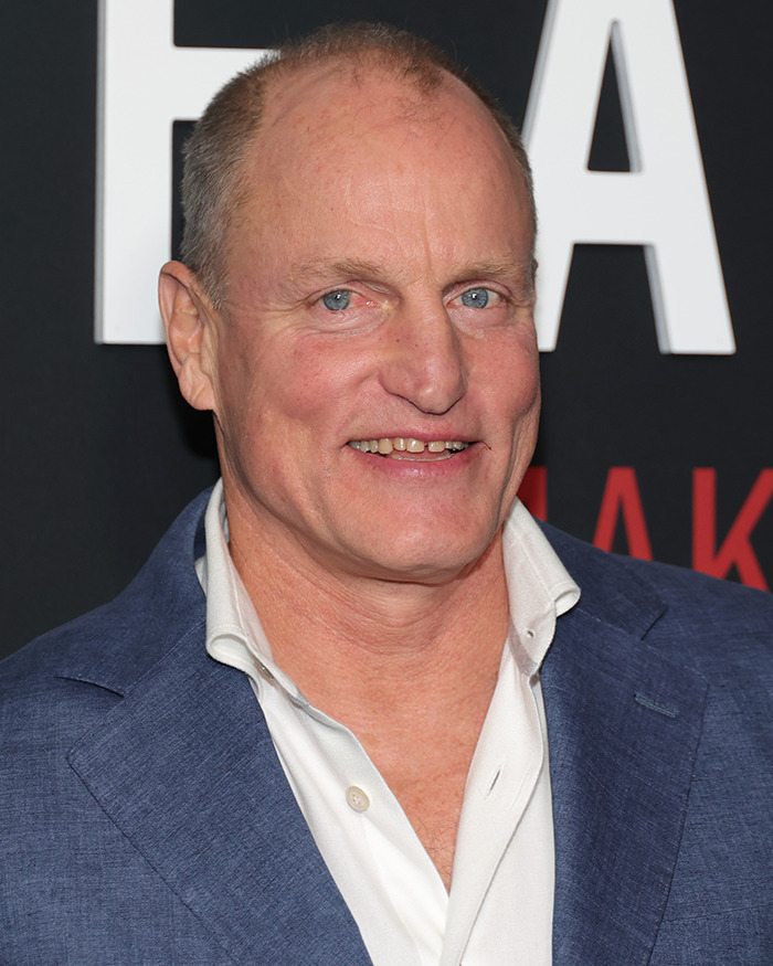 Woody Harrelson smiling at an event, wearing a blue suit jacket and white shirt, posing against a black backdrop.