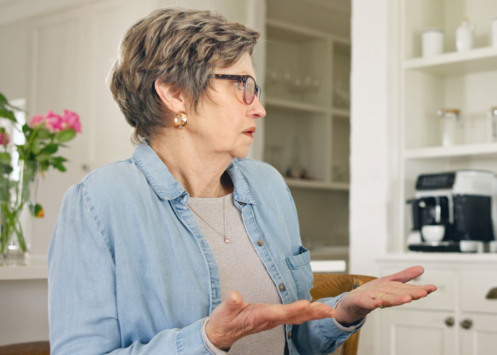 Older woman in casual clothes gesturing with hands in a kitchen, depicting tension with mother-in-law and emotional conflict.