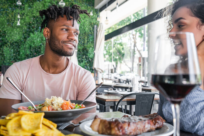 Couple on a first date at a restaurant, laughing and enjoying their meal with drinks in a bright setting.