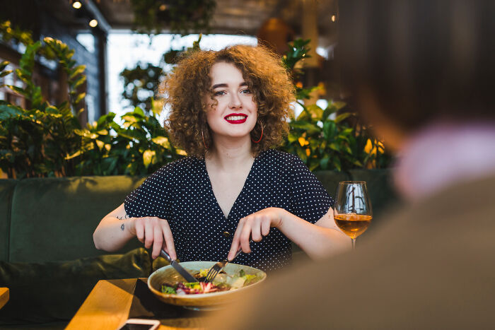 Young woman on a first date at a restaurant, smiling and eating salad with a glass of wine nearby, humorous date setting.