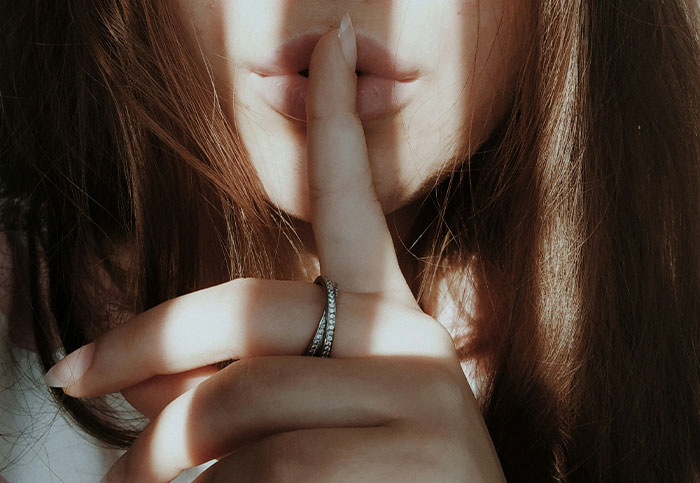 Close-up of a person with finger on lips, symbolizing silence and secrecy in someone's life being blown up unintentionally.