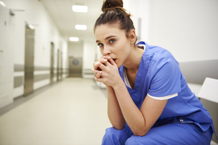 Stressed female nurse in blue scrubs sitting in hospital corridor, reflecting on haunting medical experiences.