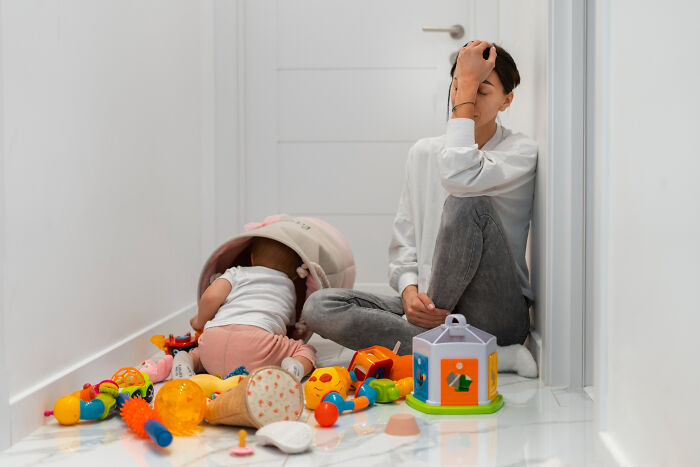 Stressed babysitter sitting on floor with baby and scattered toys, reflecting a challenging babysitting horror experience.