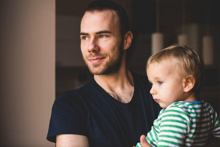 Man babysitting a toddler in a striped shirt, looking thoughtful while holding the child during a quiet moment at home.