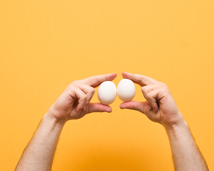 Two hands holding white eggs against a yellow background representing the concept of Scrotox cosmetic trend among men.
