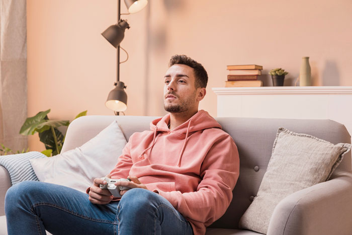 Young man in a casual pink hoodie playing video games, showing signs of laziness and entitlement at home.