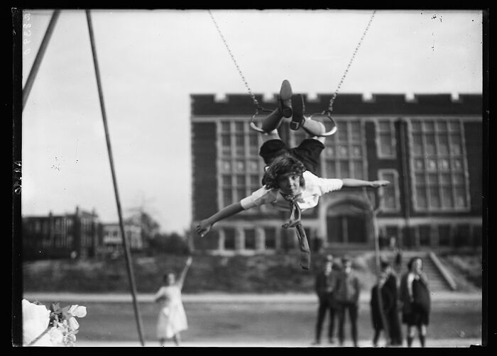 Child swinging high on vintage playground equipment with blurred school building and children in background, historic playground photo.