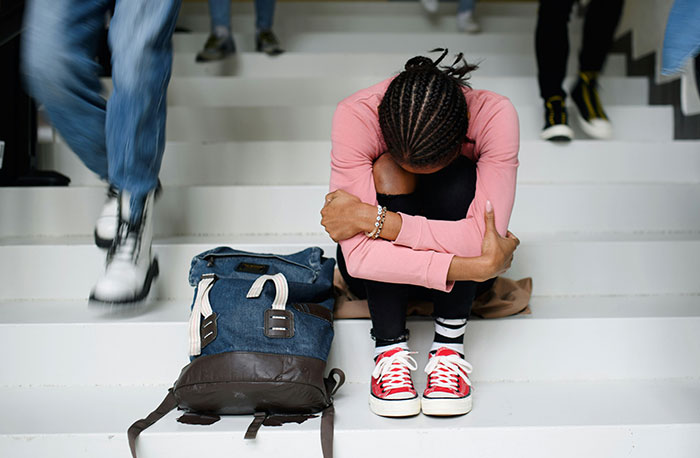 Teen girl sitting on school stairs hugging knees looking upset while other students walk by, illustrating school gossip and facts.