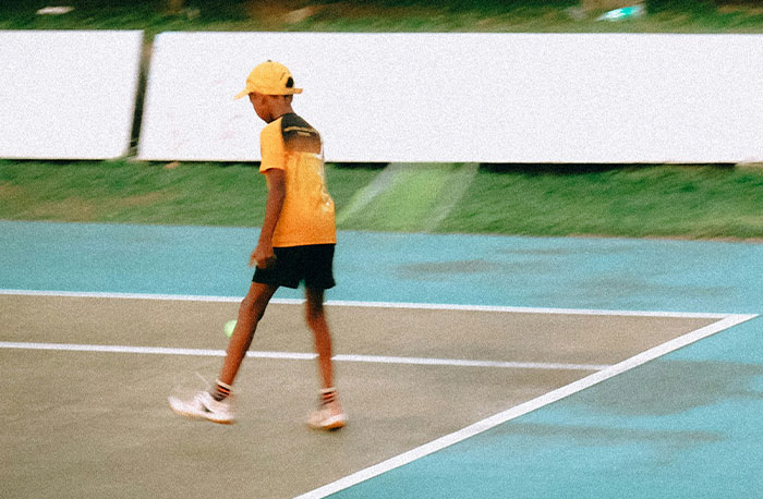 Young boy wearing a yellow cap and shirt walking alone on an outdoor sports court demonstrating school gossip themes.
