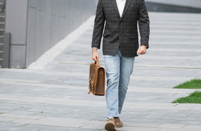 Man walking on city sidewalk wearing blazer and jeans, carrying a leather briefcase related to school gossip facts