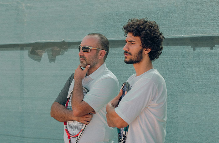 Two men holding tennis rackets, standing thoughtfully against a textured wall, representing school gossip and facts.