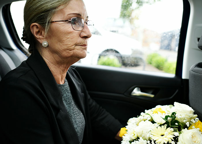 Elderly woman wearing glasses holding a bouquet of flowers in a car, reflecting on dark secrets about family members.
