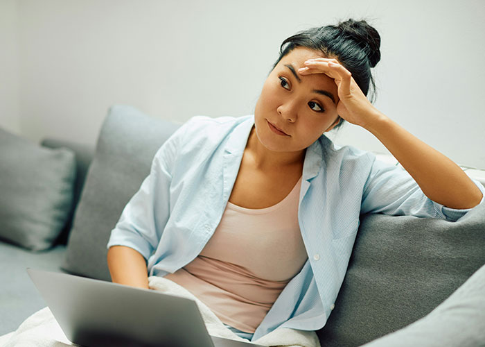 Young woman looking thoughtful and concerned on a couch with laptop, reflecting on dark secrets about family members.