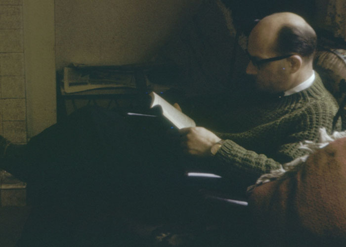Man in glasses wearing a green sweater sitting in a dim room, reading a book about dark secrets found in family members.