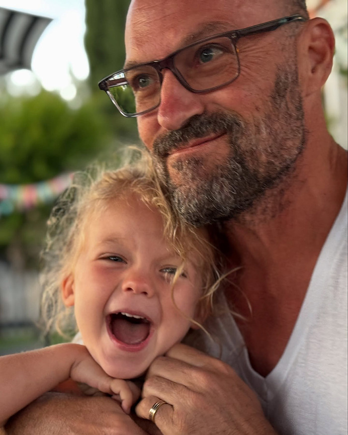 Man with glasses holding a happy young child, related to Dancing With The Stars pro defending son's feminine appearance.