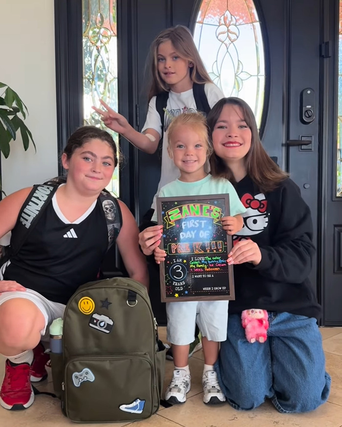 Four children posing at a front door, one holding a colorful first day of pre-kindergarten sign, showcasing a 3-year-old's appearance.