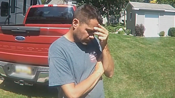Man in a gray shirt standing outside near a red truck, holding his head in distress showing a broken person reaction.