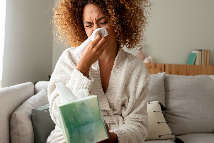 Woman in a robe sitting on a couch, blowing her nose with a tissue, depicting everyday things that humble adults.