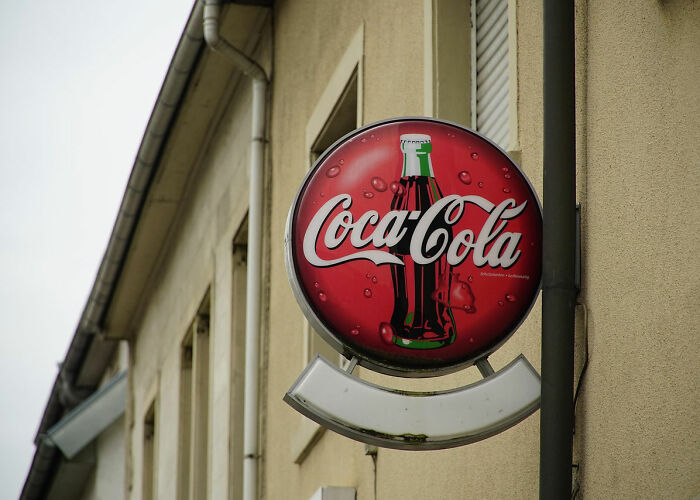 Coca-Cola round sign mounted on a beige building exterior, illustrating nostalgic rules people grew up with.
