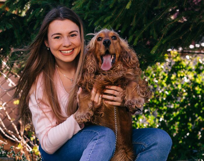 Young woman smiling while holding her therapy dog outdoors with green trees in the background on a sunny day Young woman smiling while holding her therapy dog outdoors with green trees in the background on a sunny day