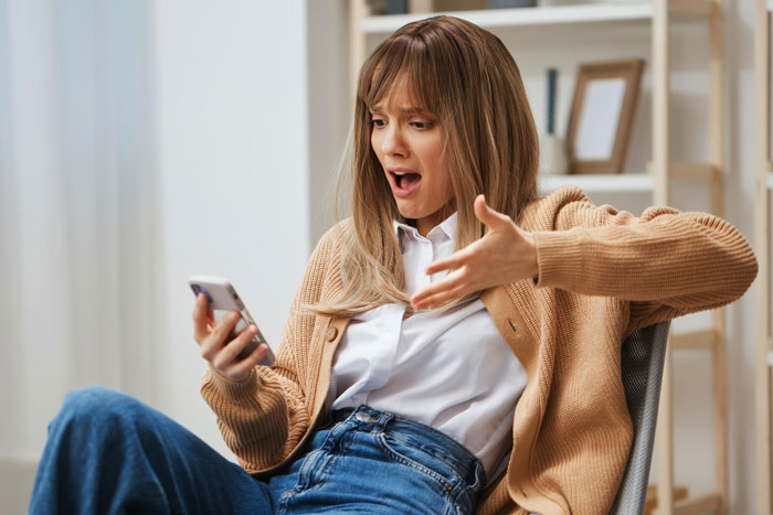 Woman in casual clothes looking shocked at her phone, feeling overshadowed during baby shower and birthday plans conflict.