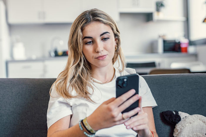 Young woman sitting on a couch looking at her phone with concern, capturing sister boyfriend relationship drama emotions.