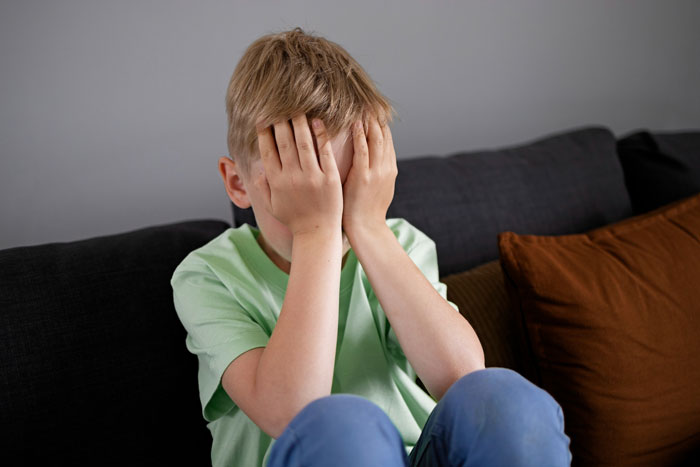Young boy sitting on couch covering face with hands, illustrating struggles with forced gender roles and family conflict. Young boy sitting on couch covering face with hands, illustrating struggles with forced gender roles and family conflict.