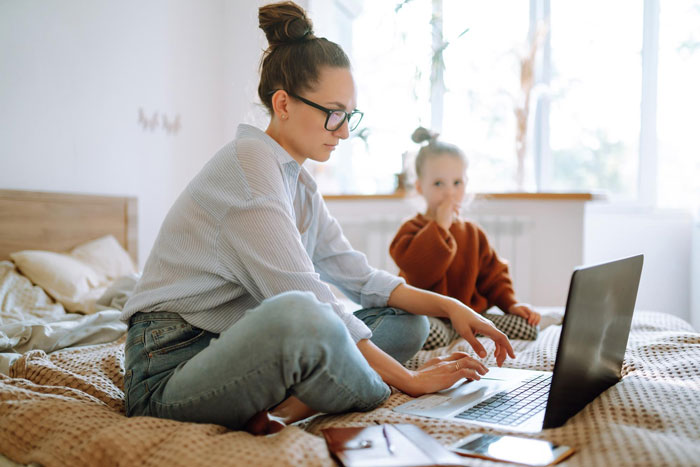 Woman in glasses working on laptop at home while child sits behind her on bed, highlighting family sitter refusal situation. Woman in glasses working on laptop at home while child sits behind her on bed, highlighting family sitter refusal situation.