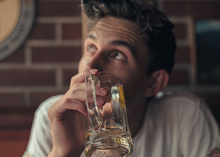 Young man holding a glass and looking up thoughtfully in a dimly lit room with a brick wall background.