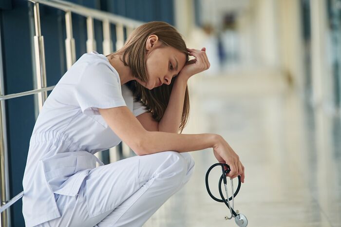 Young healthcare professional in white scrubs sitting on the floor, holding a stethoscope, appearing stressed and tired.