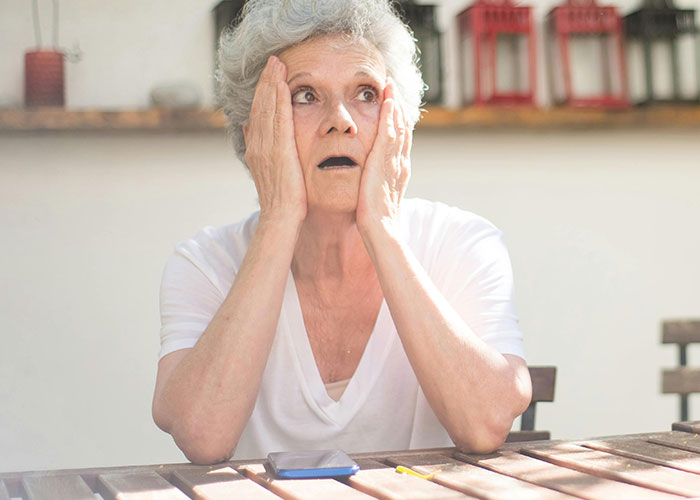 Elderly woman with a shocked expression sitting at a wooden table, reflecting on what husband thinks of her body.
