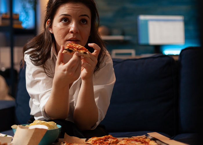 Woman sitting on couch eating pizza, reflecting on body image and emotional impact after learning husband's thoughts.