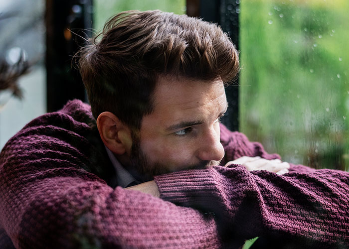 Man in a maroon sweater looking thoughtfully out a window, reflecting on feelings about body and relationship issues.