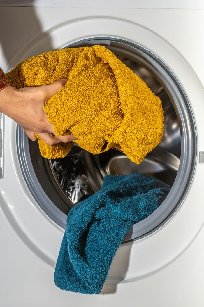 Hand placing a yellow towel into a washing machine with a blue towel inside, showing everyday things humbling adults.