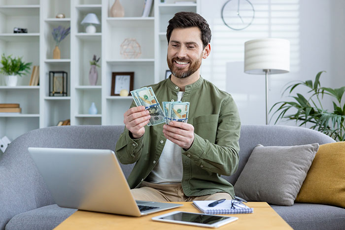 Man counting money at home, smiling while working on laptop, representing hurt working mom and ignorant husband concept.