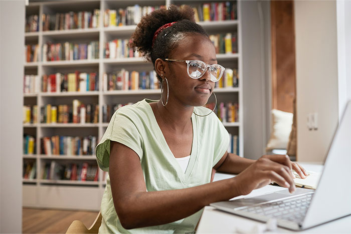 Young woman wearing glasses and hoop earrings, working on a laptop in a library, researching partner cheating cases.