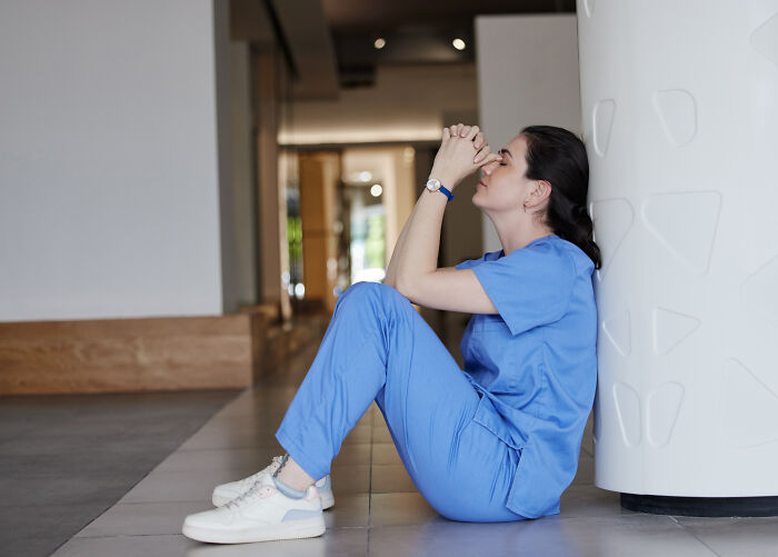 Female nurse in blue scrubs sitting on the floor, looking stressed, reflecting on haunting medical experiences.