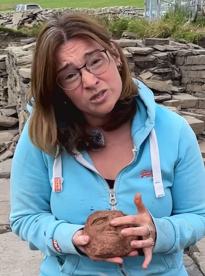 Woman in blue hoodie holding a creepy ancient head carving with a face that appears to stare back at the viewer on a farm site.