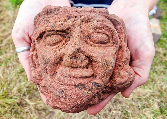 Ancient head carving with weathered features held by hands, discovered on a farm by archaeology student outdoors.