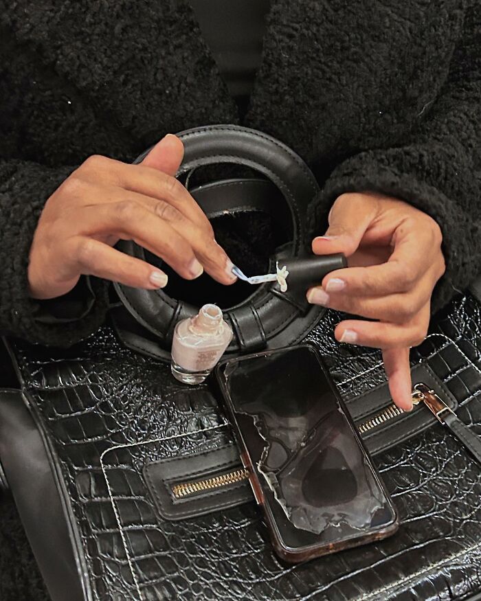 Person taking the subway in New York painting nails with nail polish while holding a black handbag and phone.
