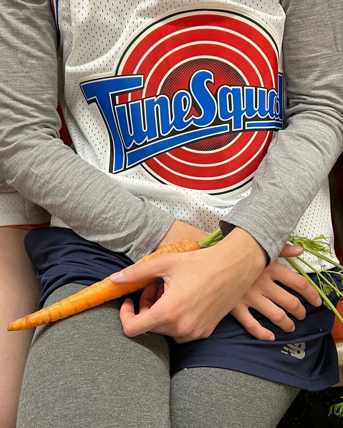 Person wearing a Tune Squad jersey holding a large fresh carrot on the subway in New York.