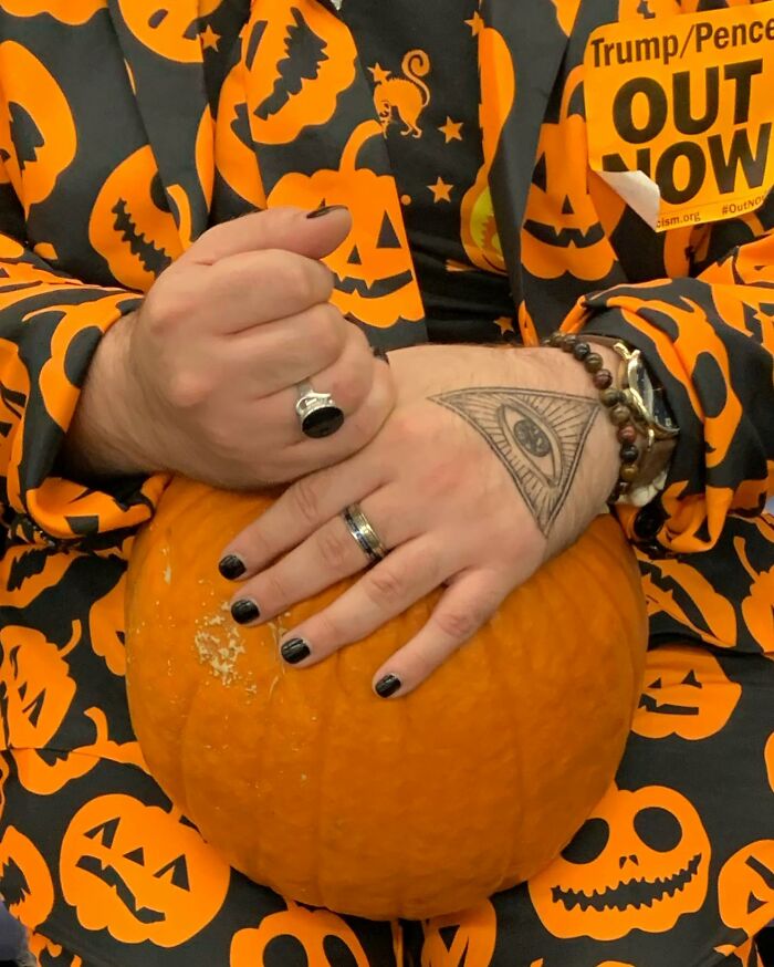 Person on New York subway holding a large pumpkin, wearing Halloween-themed clothing with jack-o-lantern patterns.