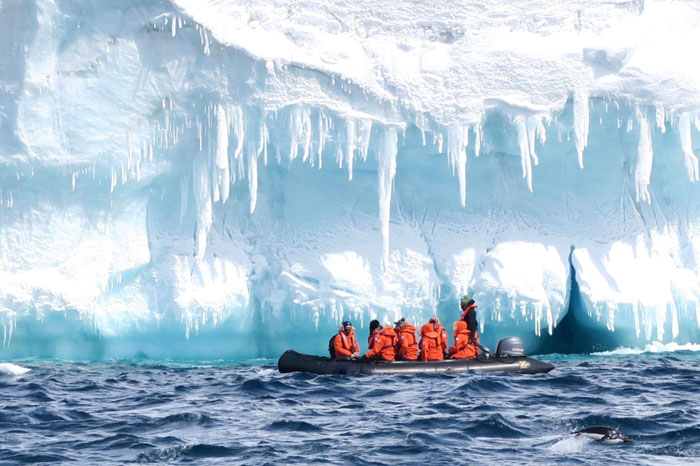 Researchers in orange jackets on a boat near large icy cliffs under a bright blue sky exploring the 1831 mystery of the sun turning blue.