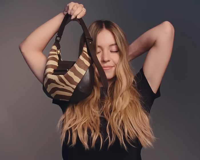 Sydney Sweeney flaunts body holding a striped Jimmy Choo bag with eyes closed against a plain background. Sydney Sweeney flaunts body holding a striped Jimmy Choo bag with eyes closed against a plain background.