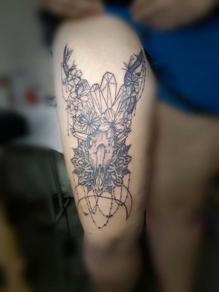 Tattoo design on thigh with intricate skull, flowers, crystals, and decorative beaded elements in black ink.