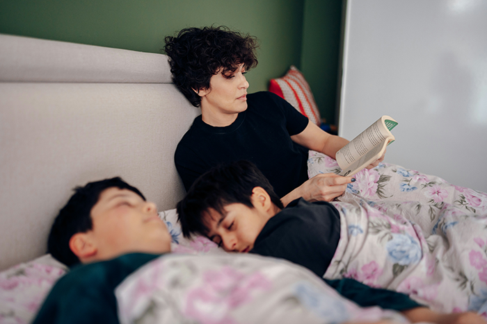 Woman reading a book in bed while two children sleep, illustrating hotel guests disturbing neighbors with late-night ruckus. Woman reading a book in bed while two children sleep, illustrating hotel guests disturbing neighbors with late-night ruckus.