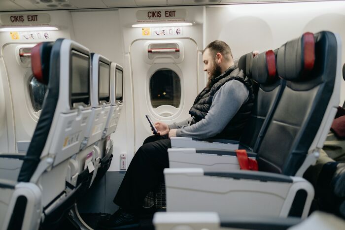 Man seated alone in airplane cabin near exit row, using smartphone, illustrating people who got fired for careless mistakes.